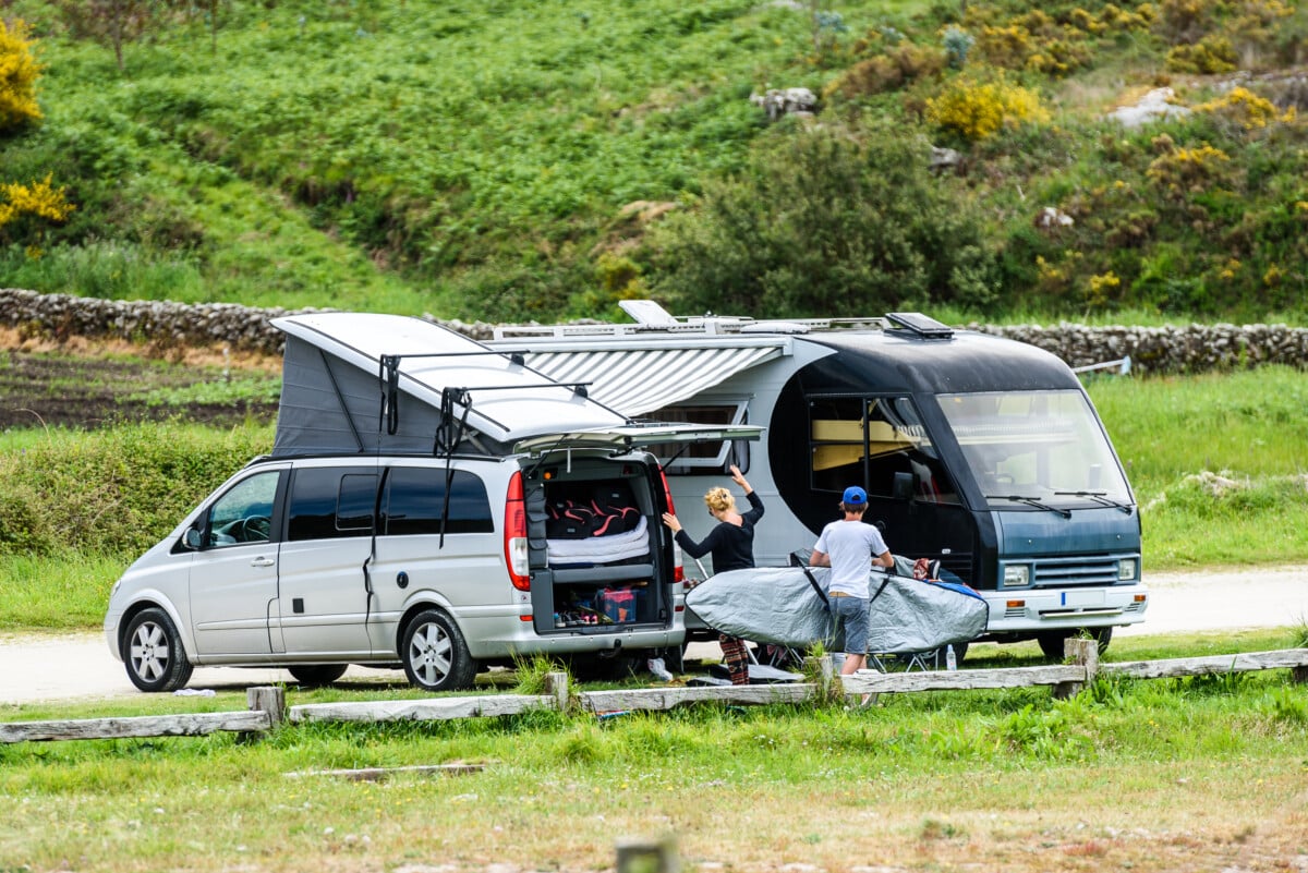 Motorhome RV and campervan are parked on a beach. Surfers on vacation are packing surfboards or boards after a day of wave surfing.