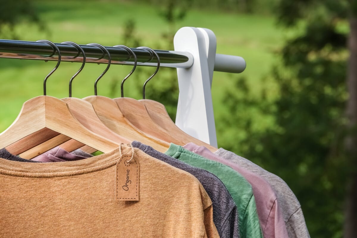 Organic clothes. Natural colored t-shirts hanging on wooden hangers in a row. Eco textile tag. Green forest, nature in background.