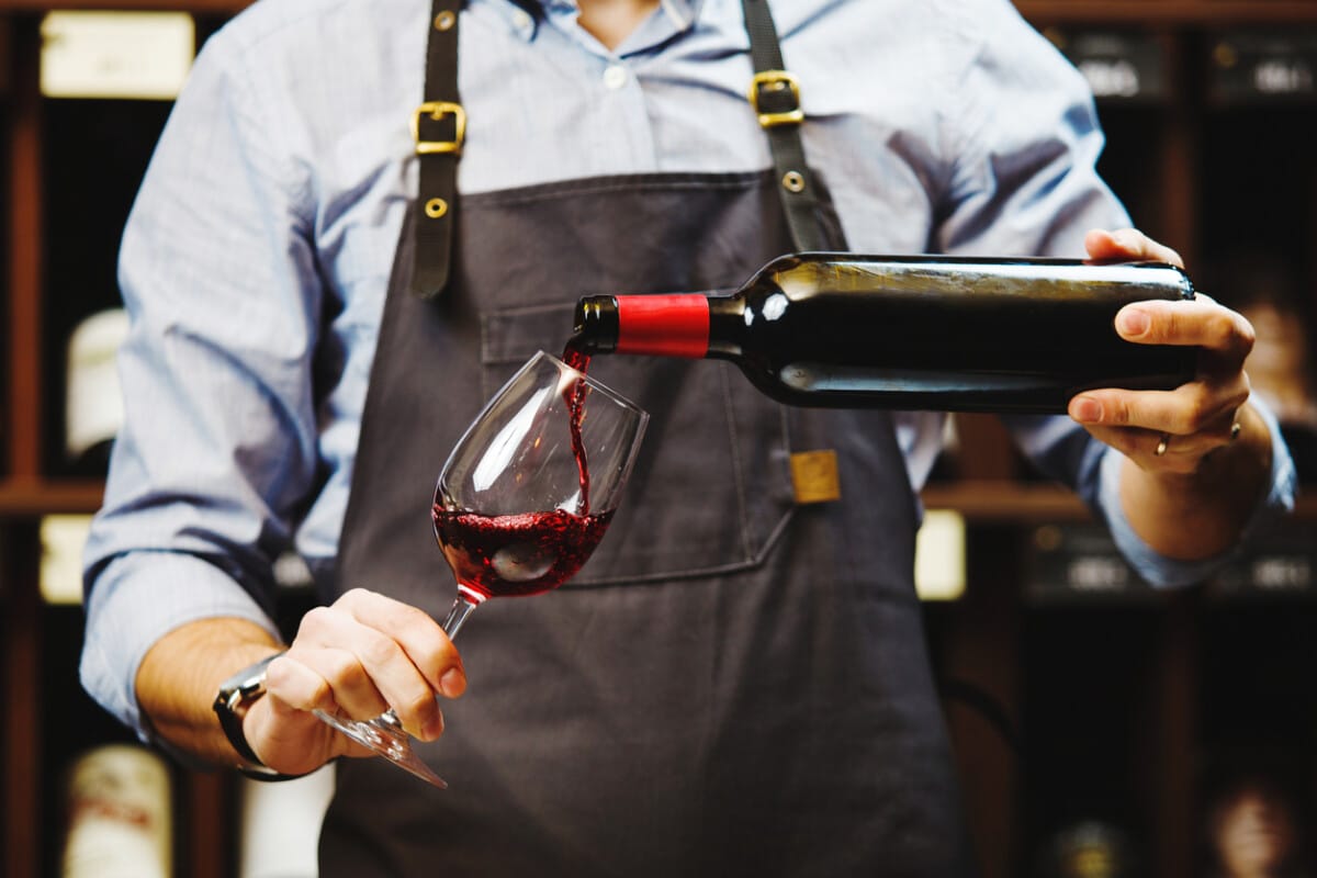 Male sommelier pouring red wine into long-stemmed wineglasses. Waiter with bottle of alcohol beverage. Bartender at bar counter pour elite drink into long-stemmed glass