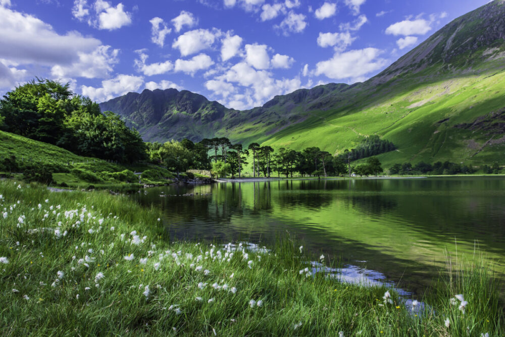 Idyllic scenery of English Lake District in springtime.Trees and grass growing on lakeshore and green hill reflecting in lake water. Sunlight kissing mountain slope in background.Majestic landscape scene. This is a perfect destination for UK family adventures.