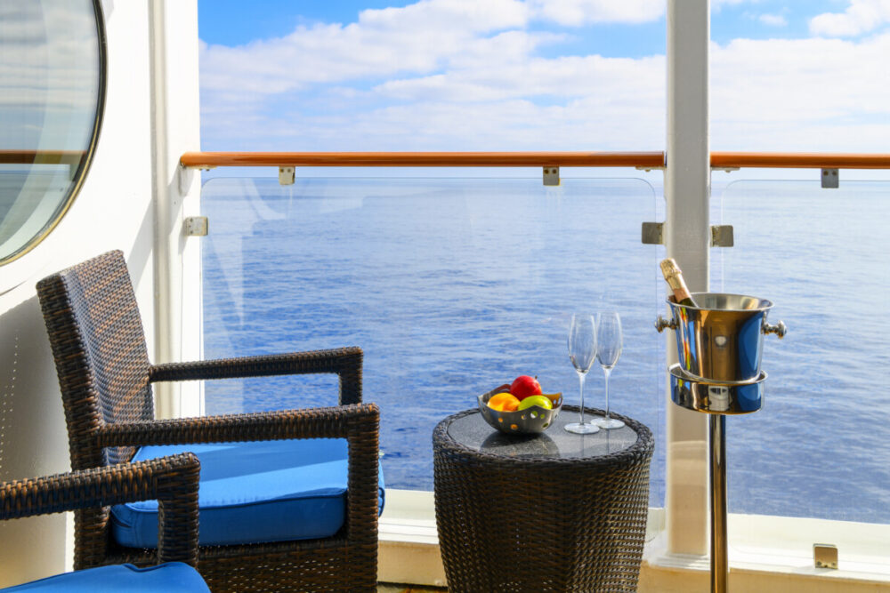 A bowl of fresh fruit, a bottle of champagne in an ice bucket, and two champagne glasses on an oceanfront balcony veranda on a sunny cruise ship.