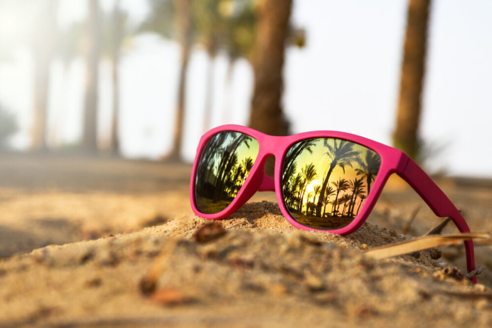 Elevate Your Look: Perfect Eyewear Colors for Every Destination 2 Pink modern sunglasses on the beach sand with palm trees and sunset reflection