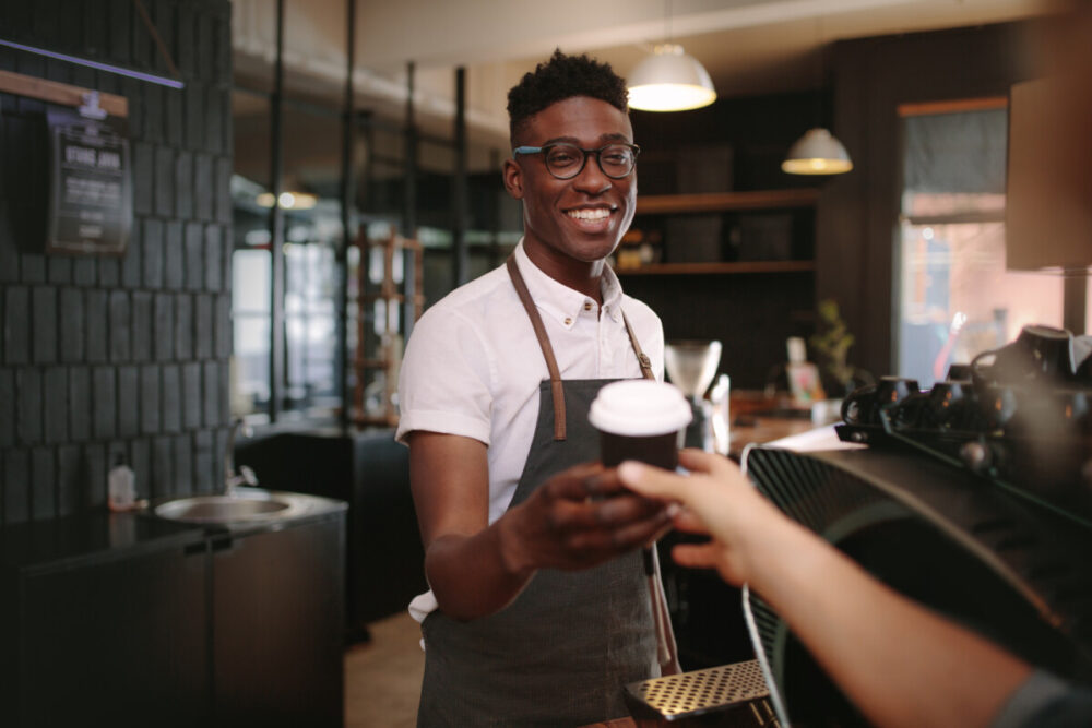 How to Plan a Culinary Trip Around the World 1 Coffee shop owner handing over a sealed coffee cup to a customer. Man serving customer with a smile at a coffee shop.