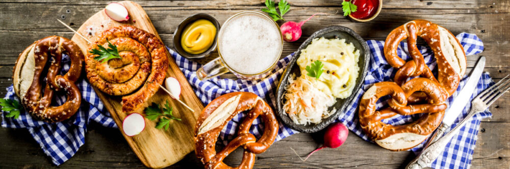 How to Make the Most of Munich Oktoberfest 2 Beer Fest food menu, bavarian sausages with pretzels, mashed potato, sauerkraut, beer bottle and mug old rustic wooden background, copy space above banner format