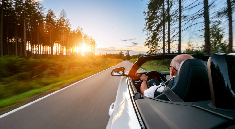 Mature Adult man is driving with convertable car in sunny nature on a bright summer day. wide angle pursuit shot with high speed motion blur. Car finance terms before travel. Long trip travel tips.
