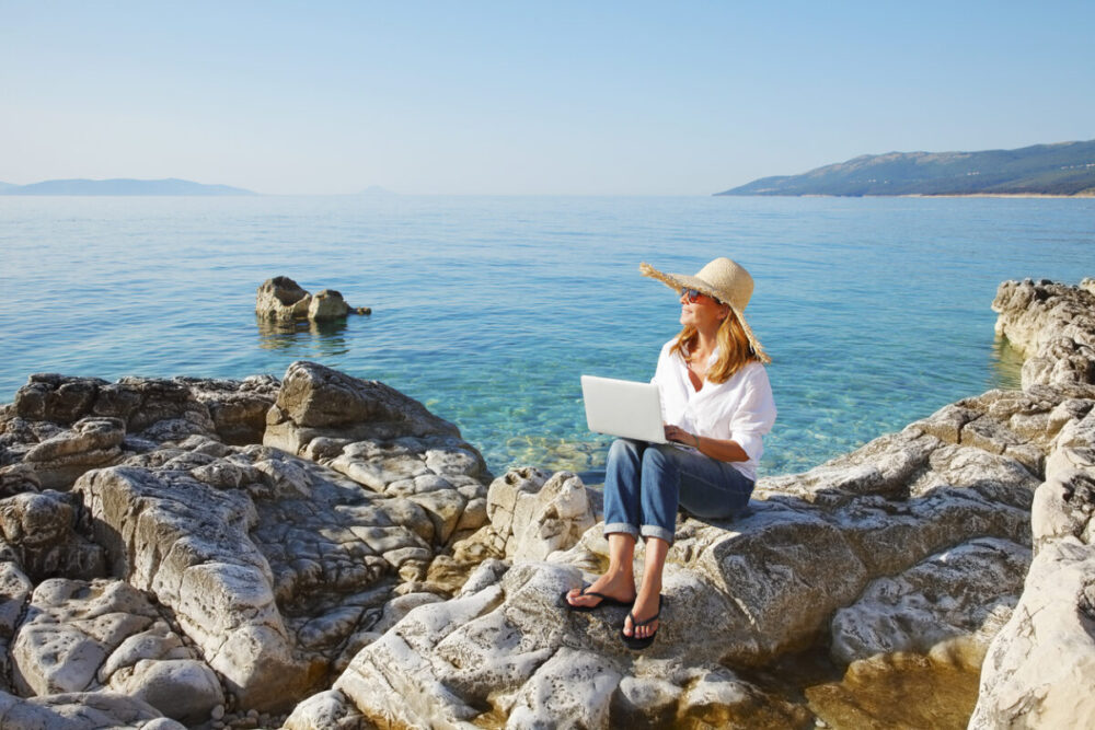 9 Productivity Tips for Digital Nomad Writers 1 Full length portrait of casual businesswoman working by the sea. Middle age female sitting on the rock and typing on her laptop.