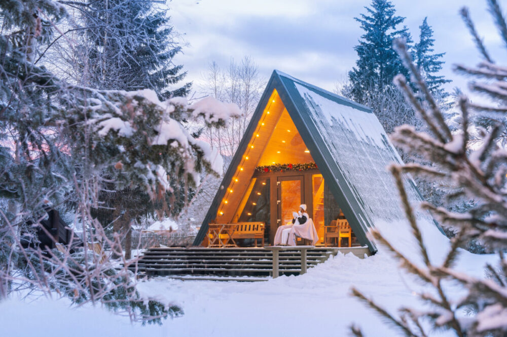 What to Pack for a Cozy Cabin Weekend in Fall and Winter 1 warm drink on the porch of an A-frame cabin in a serene winter forest during the early morning light. A woman drinks coffee or tea in a quiet evening in a house near the forest in the late evening