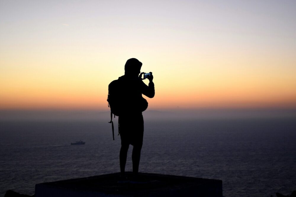 How to Shoot Magazine‑Worthy Travel Images With Your Smartphone 3 silhouette photography of person standing in front of calm body of water during golden hourPerson taking a picture of the sunset