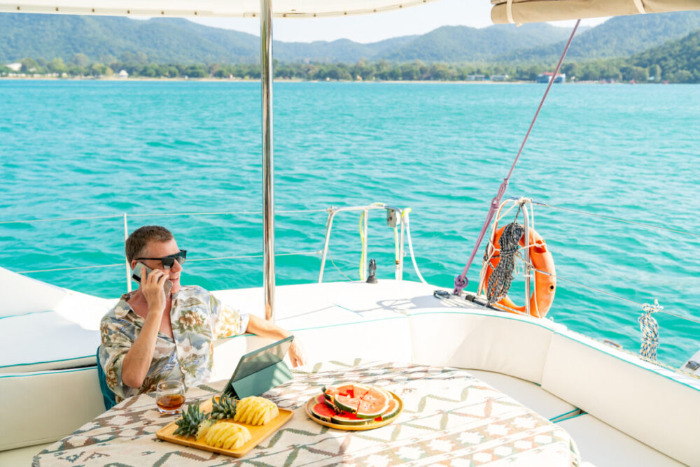 The Sophisticated Nomad: Curating Digital Safety for Global Adventures 1 Workation on summer beach holiday vacation. Caucasian businessman working on digital tablet and speaking on mobile phone for online corporate business on luxury catamaran boat yacht sailing in ocean.