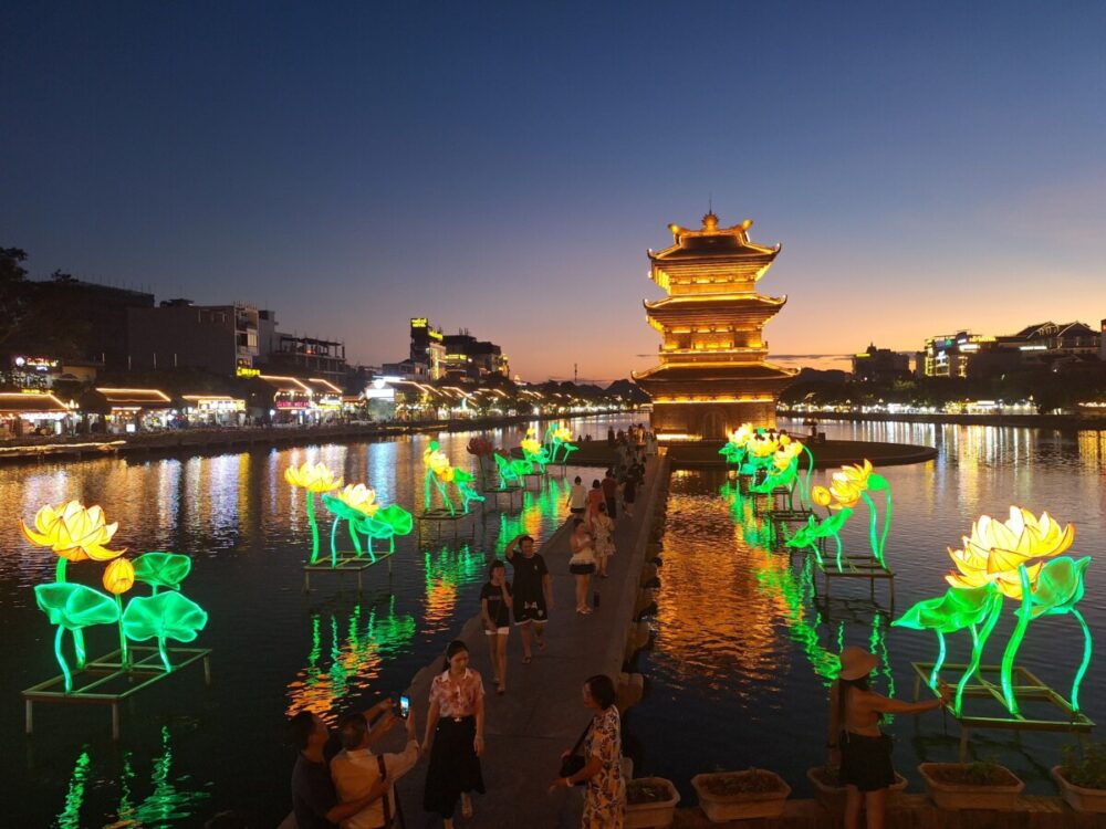 Beyond the Karsts: How Pullman Ninh Binh Unlocks the Real Ninh Binh 8 An illuminated multi-tiered pagoda stands at twilight beside a calm lake decorated with large, glowing artificial lotus flowers. Buildings with bright lights line the opposite shore, reflecting vibrant colors onto the water's surface beneath a sunset sky fading from orange to blue.