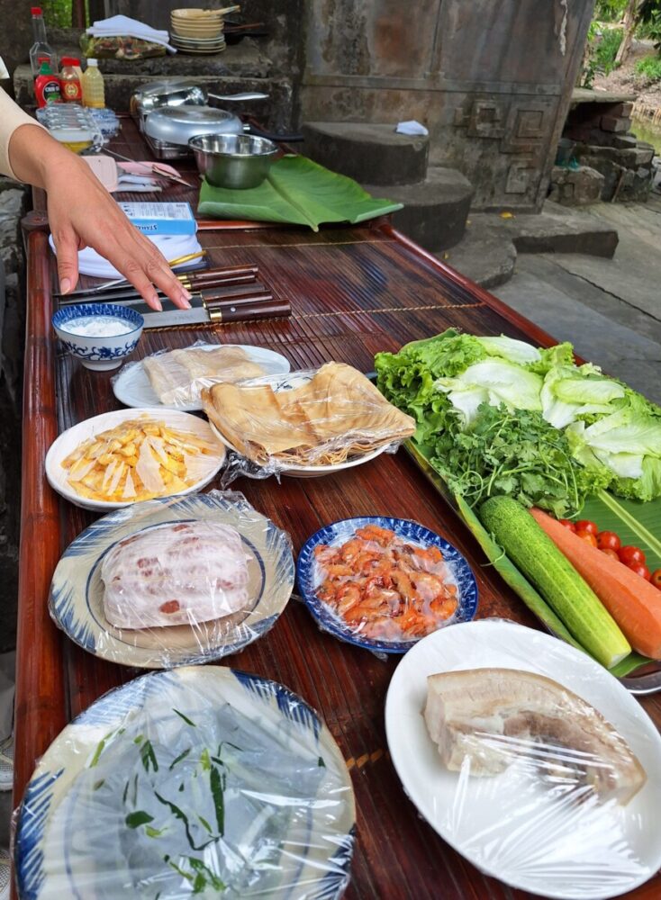 Beyond the Karsts: How Pullman Ninh Binh Unlocks the Real Ninh Binh 6 A wooden table is spread with fresh ingredients for a Vietnamese meal, including sliced pork belly, shrimp, rice paper sheets, herbs, and fresh vegetables wrapped in plastic wrap.