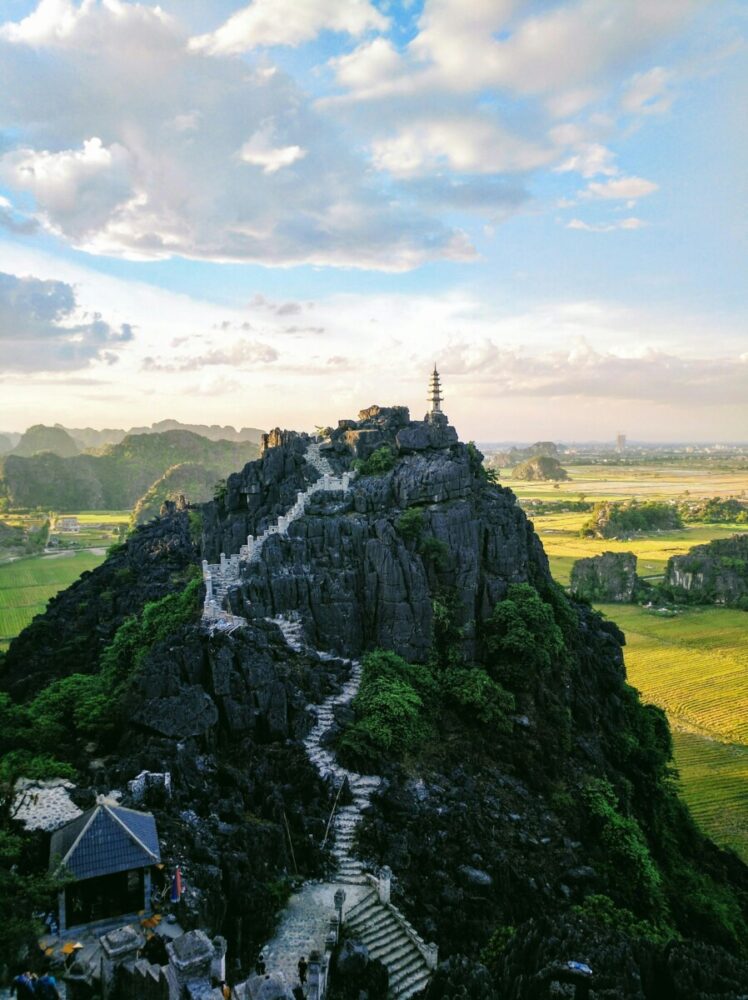 Beyond the Karsts: How Pullman Ninh Binh Unlocks the Real Ninh Binh 5 An aerial view shows a winding stone staircase climbing up a steep, rocky mountain peak toward a small pagoda at the summit, overlooking a vast landscape of green and yellow rice fields under a cloudy blue sky.