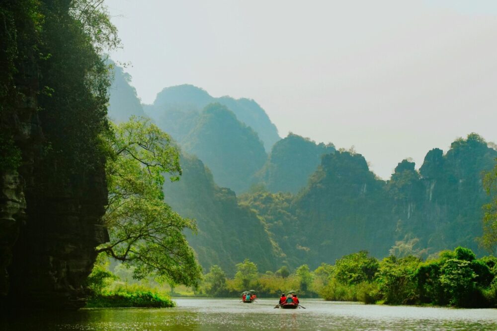 Beyond the Karsts: How Pullman Ninh Binh Unlocks the Real Ninh Binh 3 A scenic river flows between towering limestone mountains covered in lush green vegetation under a hazy sky, with small paddle boats carrying passengers in the distance.