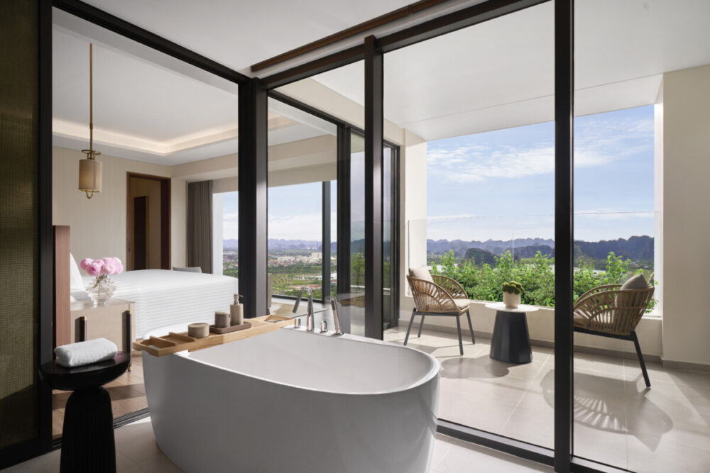 Beyond the Karsts: How Pullman Ninh Binh Unlocks the Real Ninh Binh 9 A modern luxury hotel suite featuring an open-concept bathroom with a freestanding white bathtub. The outdoor view showcases a lush green landscape with distant mountains under a bright sky.