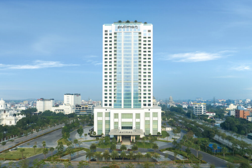 Beyond the Karsts: How Pullman Ninh Binh Unlocks the Real Ninh Binh 2 An aerial wide shot of the modern, white high-rise Pullman Ninh Binh hotel tower standing prominently against a bright blue sky with light, scattered clouds.