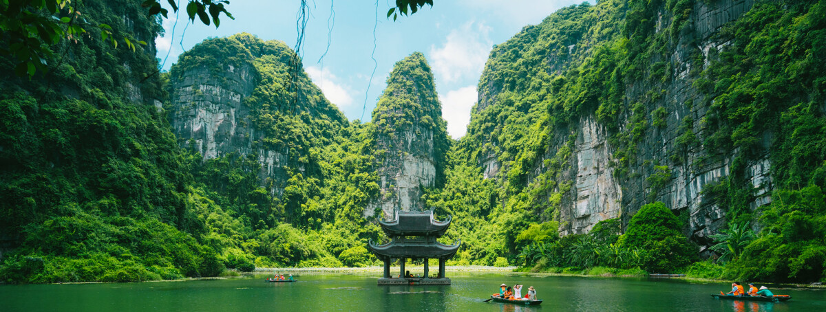An ancient stone pavilion sits on a calm green river surrounded by towering limestone mountains covered in lush green vegetation, with small wooden boats carrying tourists in Vietnam's Tràng An Scenic Landscape Complex.