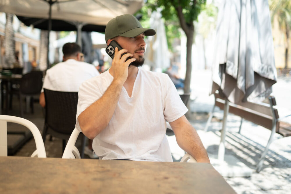 How to Choose the Best eSIM for Europe Travel 1 Young bearded man wearing a cap is sitting at a table in an outdoor cafe, talking on his mobile phone