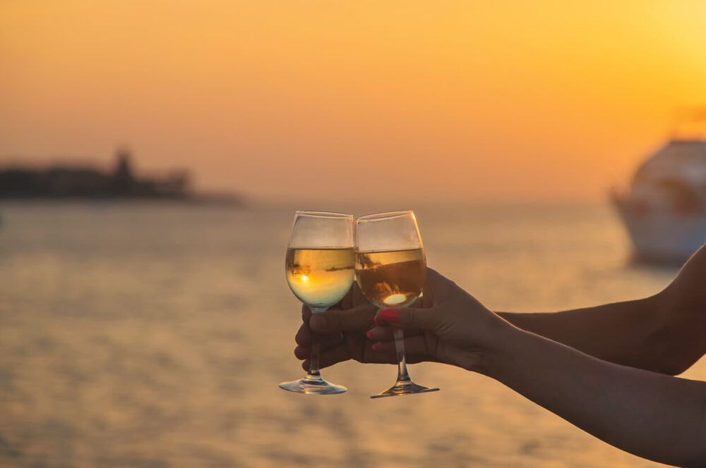 Two glasses of white wine resting on the railing of a ship overlooking the ocean at sunset