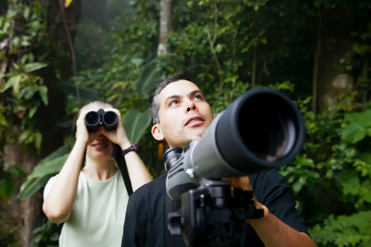 A woman and man in the jungle using binoculars and a telescope.