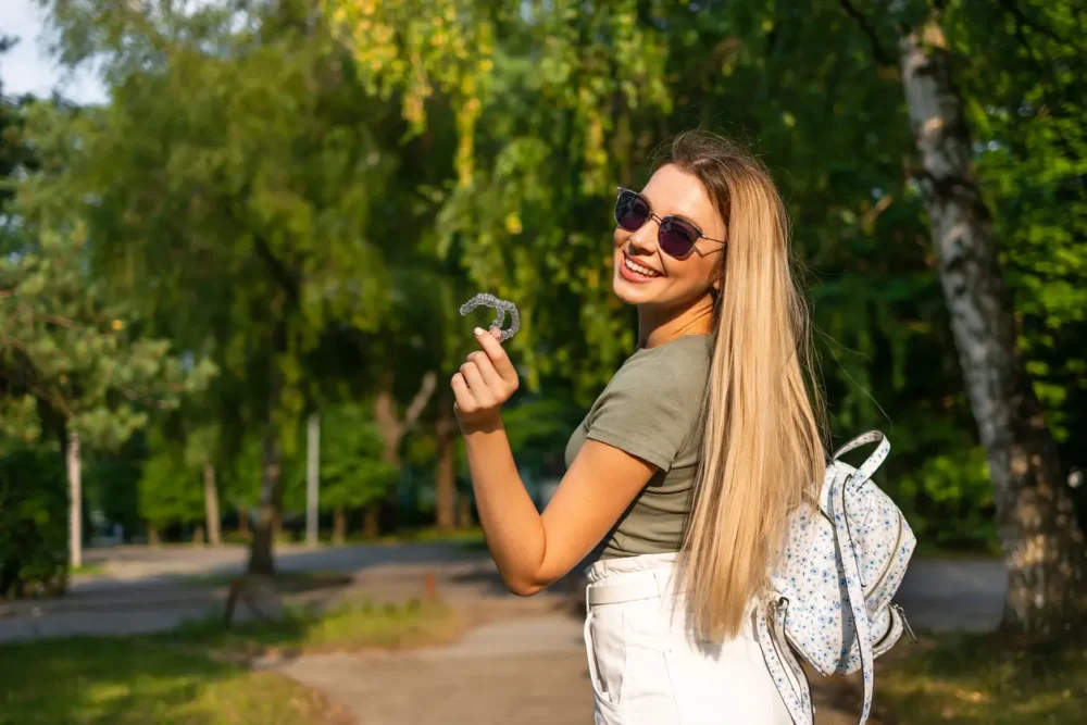 How to Manage Traveling With Clear Aligners Like a Pro 2 Smiling woman with invisible aligner in her hand, backpack on her shoulder, walking down a wooded path